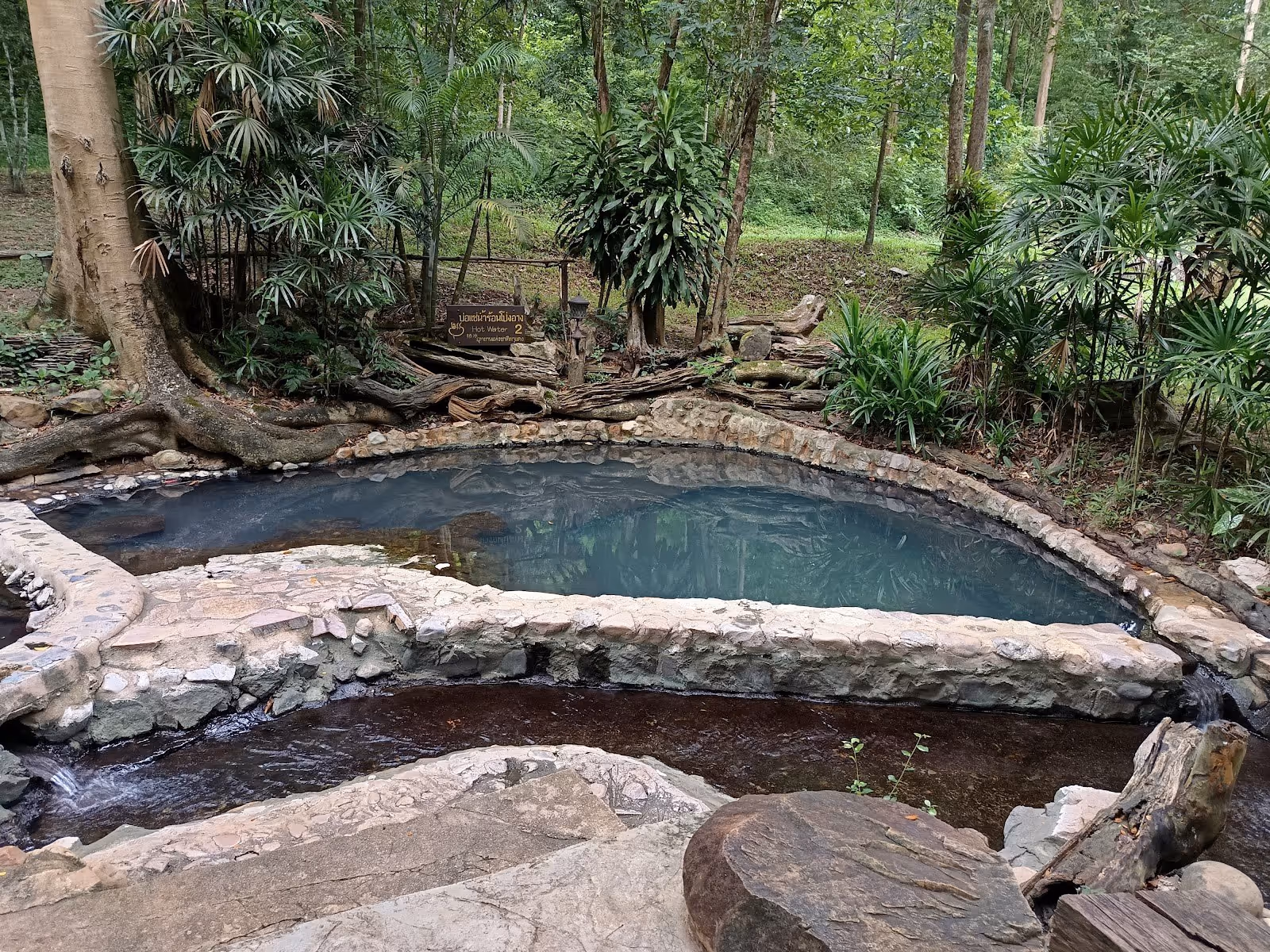 Stone-bordered soaking pool filled with clear water surrounded by trees at Pong Arng Hot Springs near Chiang Dao, Nan, Thailand.
