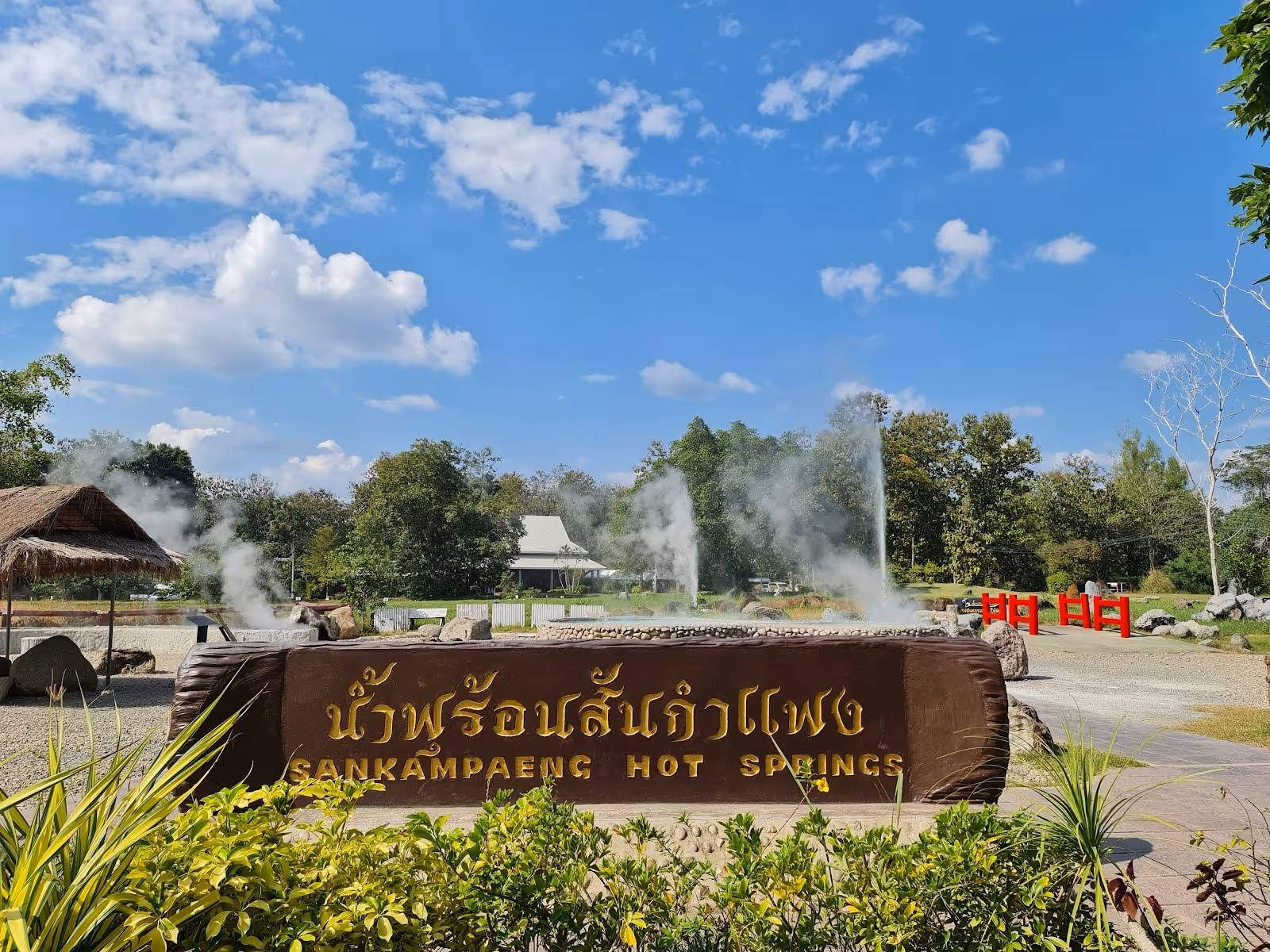 Wooden entrance sign for San Kamphaeng Hot Springs with steaming geysers and trees on a sunny day near Chiang Mai, Thailand.