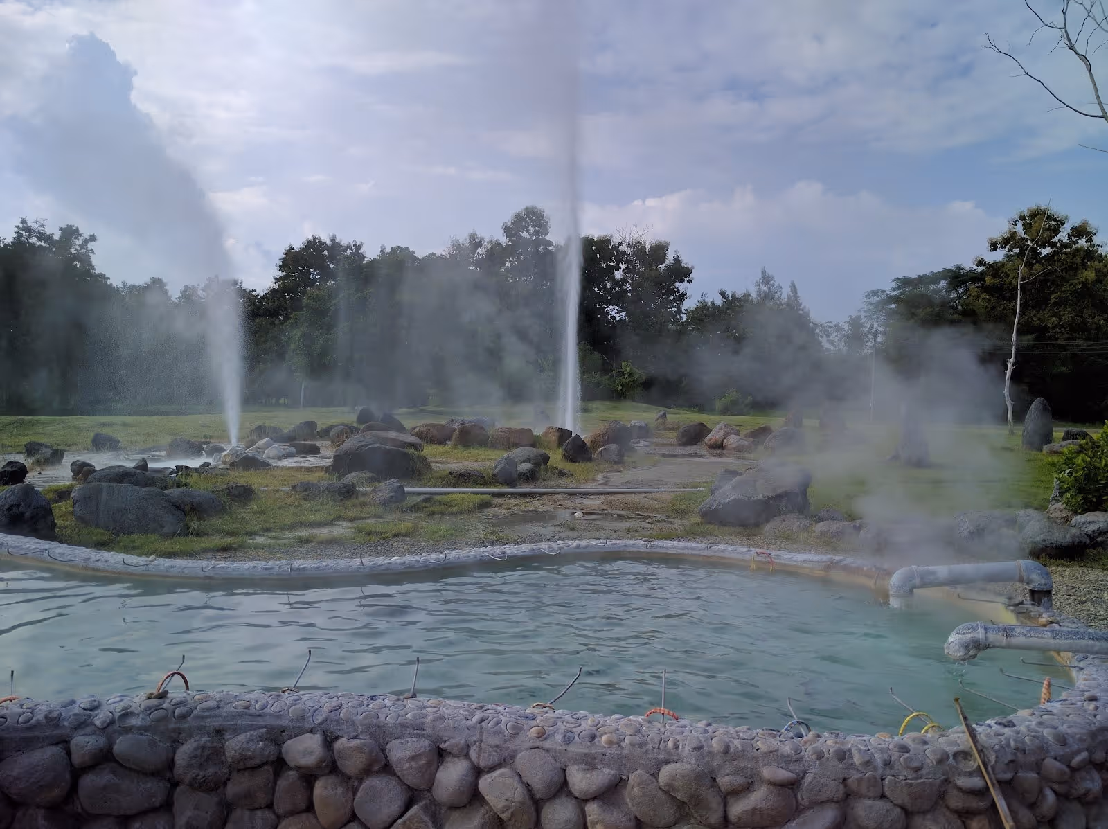 Several geysers releasing steam around a mineral water pool with stones at San Kamphaeng Hot Springs in Nan, Thailand.