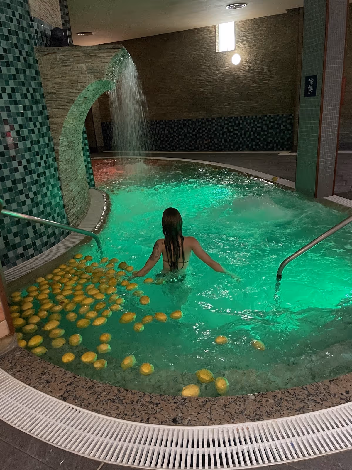 Person soaking in Balneario de Archena indoor hot spring pool with green lighting and curved stone waterfall in Murcia, Spain.