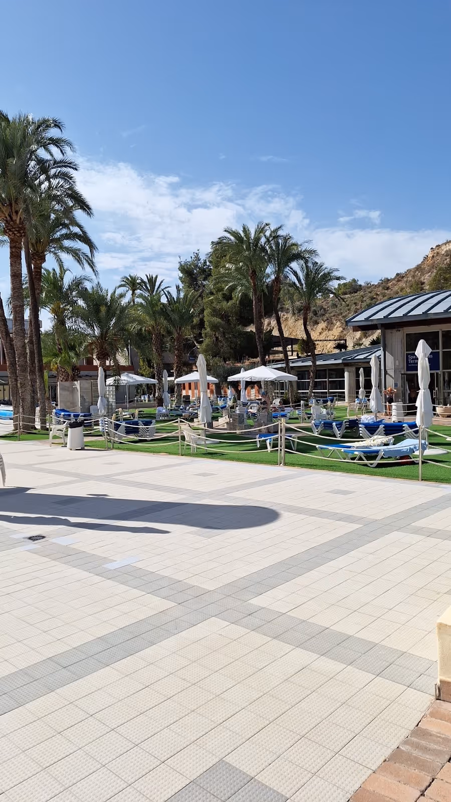 Palm trees and lounge chairs around outdoor mineral pools under a blue sky at Balneario de Archena near Murcia, Spain.
