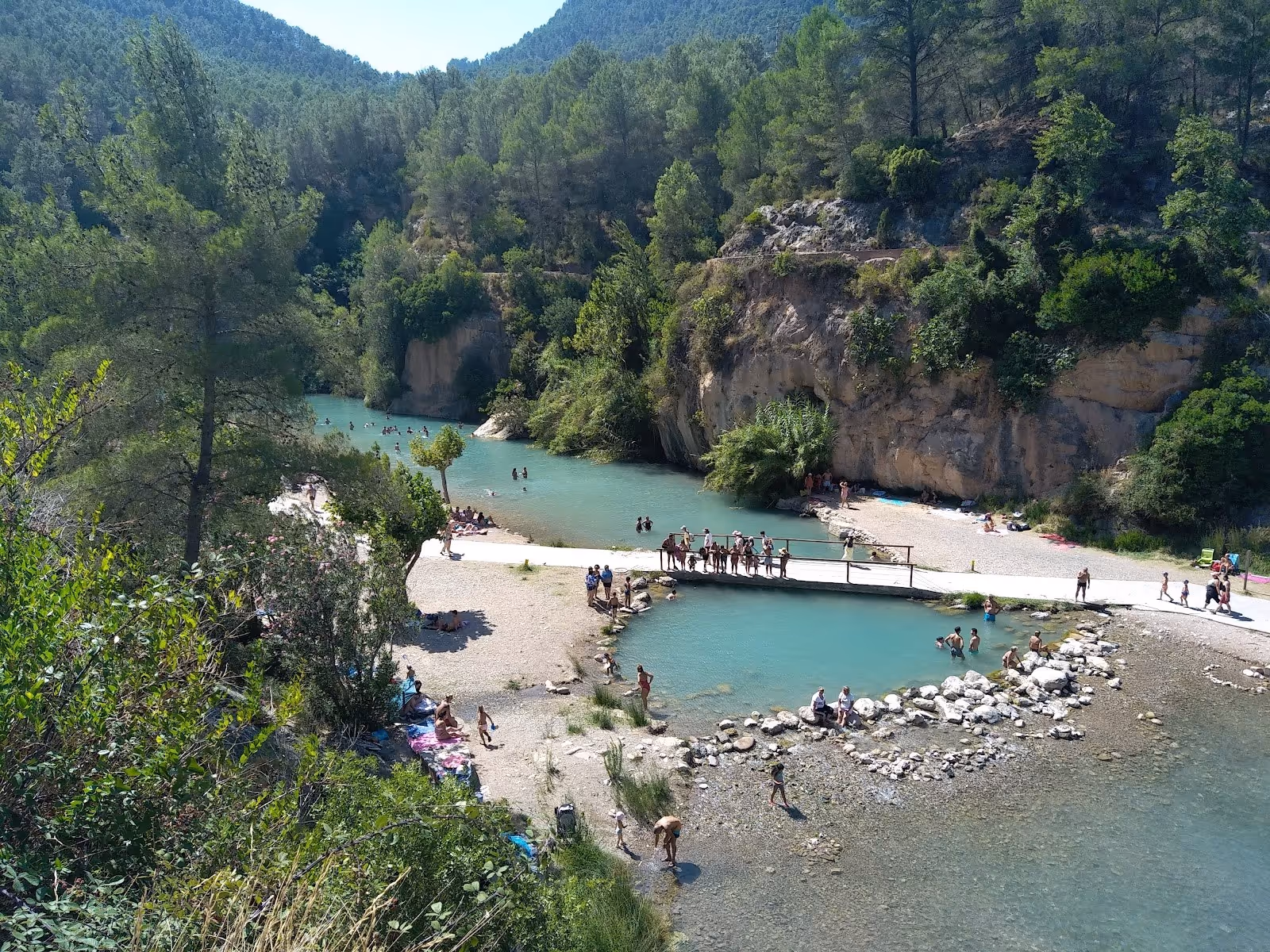 Natural outdoor hot spring pool surrounded by green trees and rocky cliffs with visitors bathing and relaxing in Montanejos, Spain.
