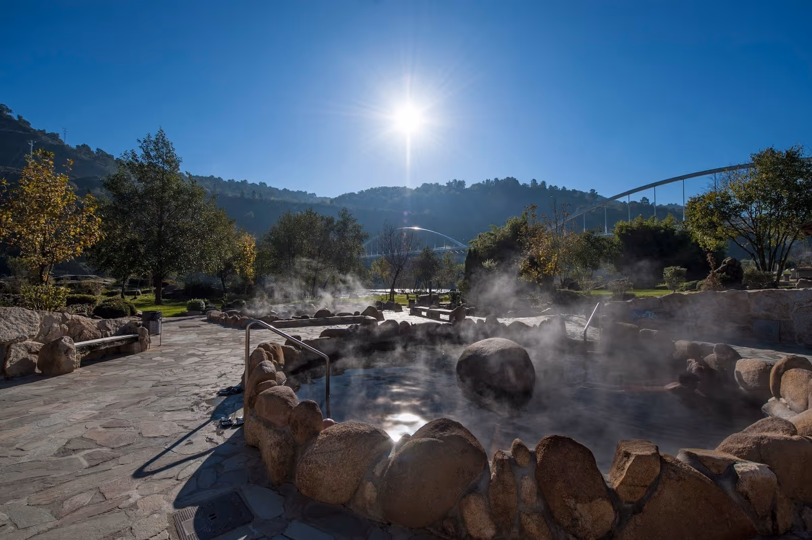 Steaming circular hot spring pool surrounded by large rocks with sun shining and a bridge in the background in Ourense Spain.