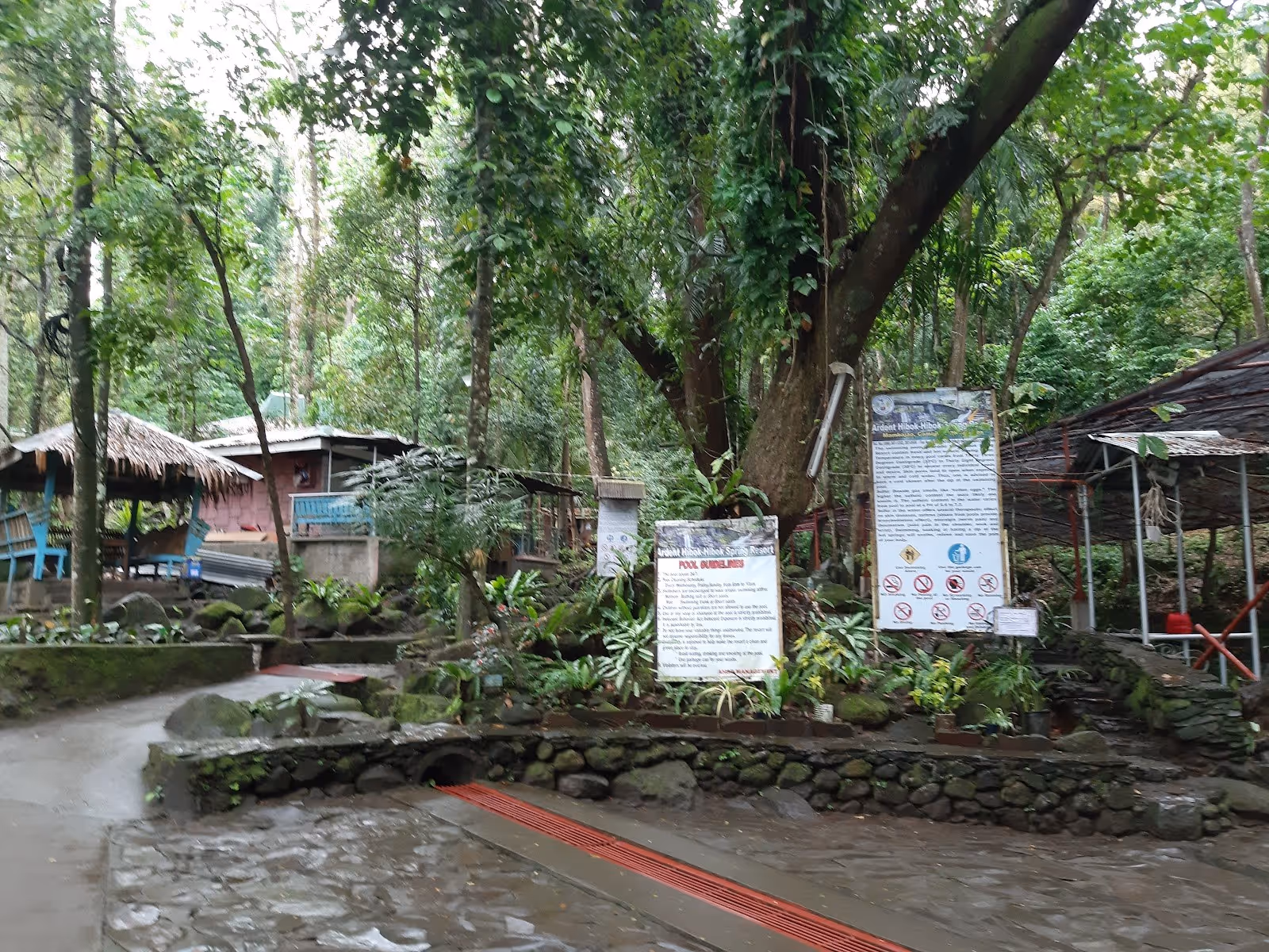Pathway and signs near shaded seating areas and huts surrounded by trees at Ardent Hot Spring in Mambajao.