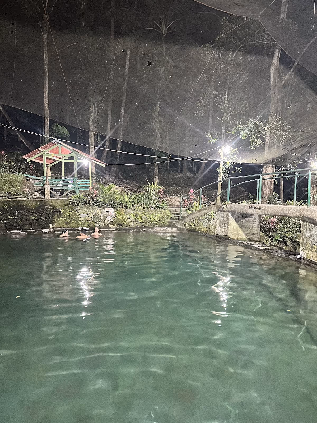 Outdoor hot spring pool at Ardent Hot Spring in Mambajao with stone edges and a small covered seating area at night.