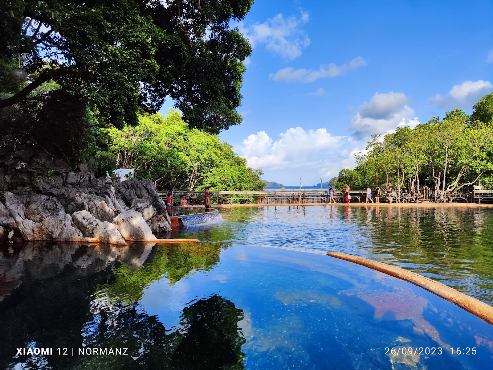 People relaxing in a calm outdoor mineral hot spring pool with rocky edges and lush green trees at Maquinit Hot Spring in Coron.