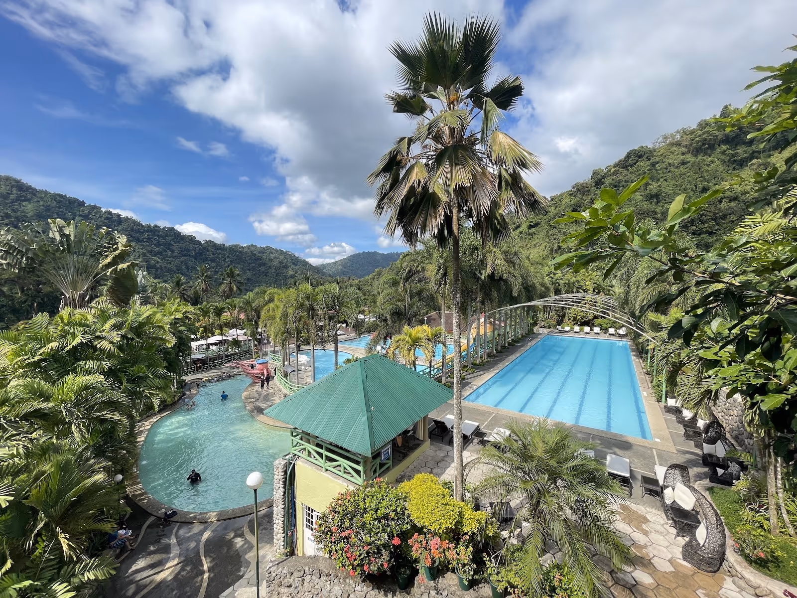 Aerial view showing multiple crystal blue pools, palm trees, and tropical plants with mountains in the distance at Palm Grove Hot Springs i.