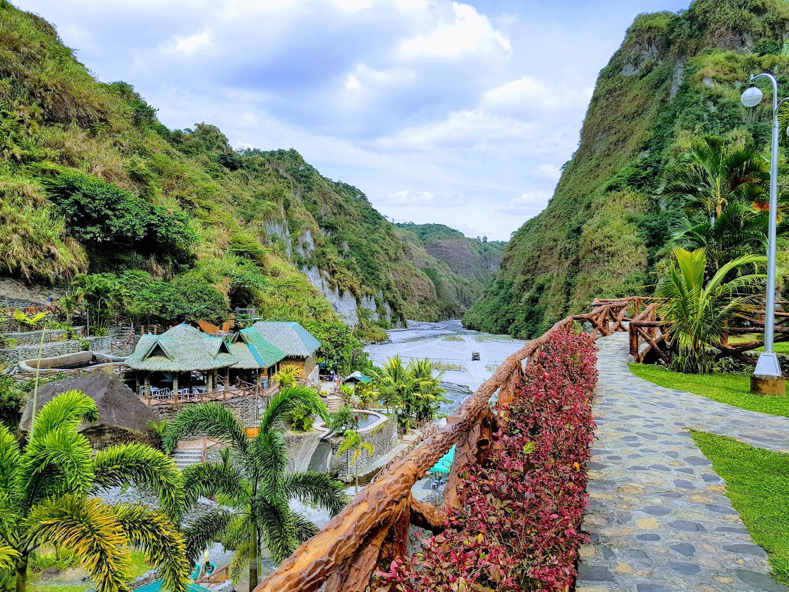 Stone pathway with wooden railing along red shrubs overlooking green hills and traditional huts near Puning Hot Spring in Angeles City Phil.