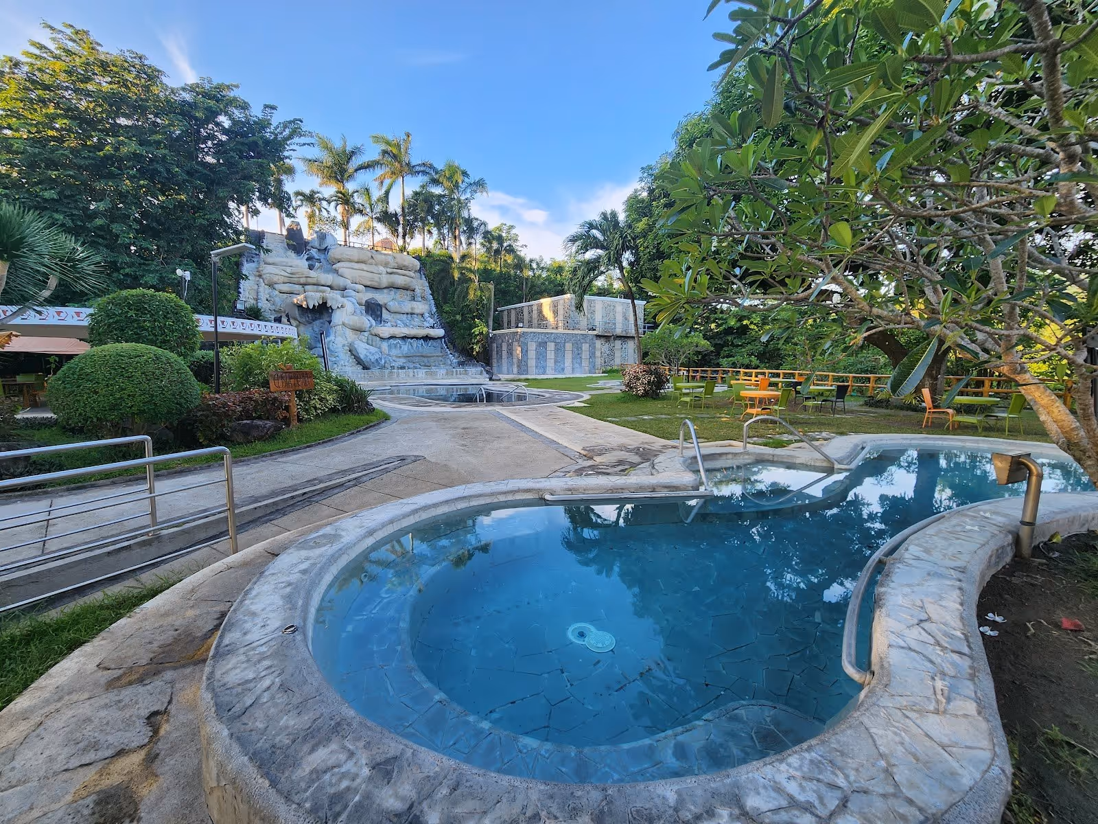 Round stone hot spring pool surrounded by green trees and chairs with view of the waterfall at Sol Y Viento in Calamba City Philippines.