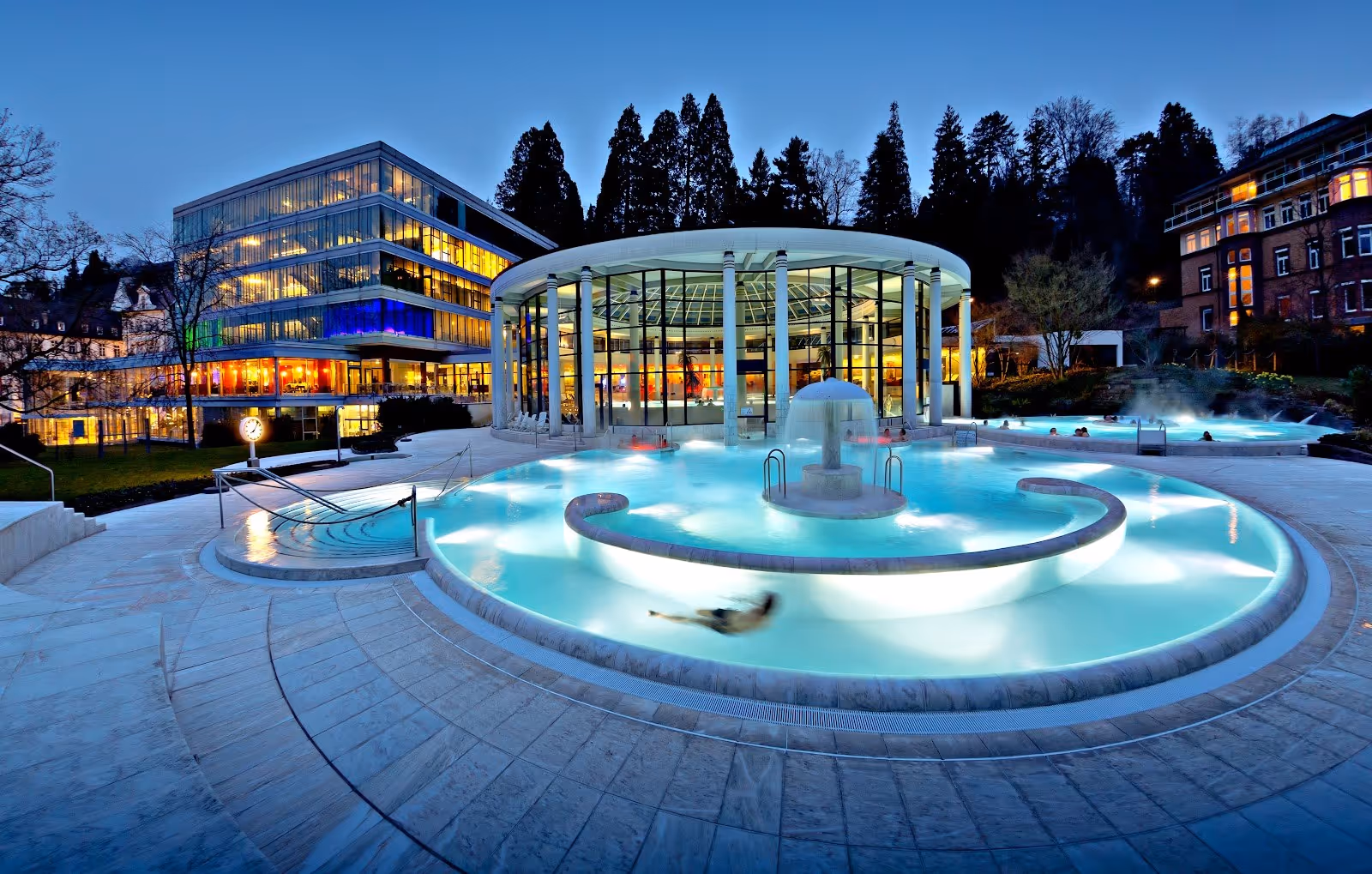 Outdoor hot spring pool at Caracalla Therme in Baden-Baden Germany with steam rising and illuminated fountain at dusk.
