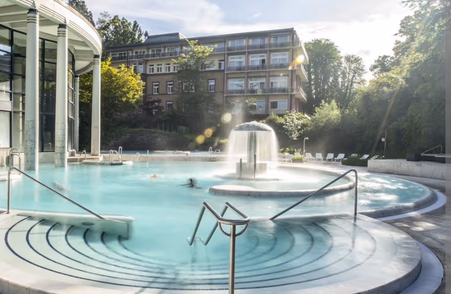 Sunlit outdoor mineral pool at Caracalla Therme with a central water feature and curved steps leading into the water.