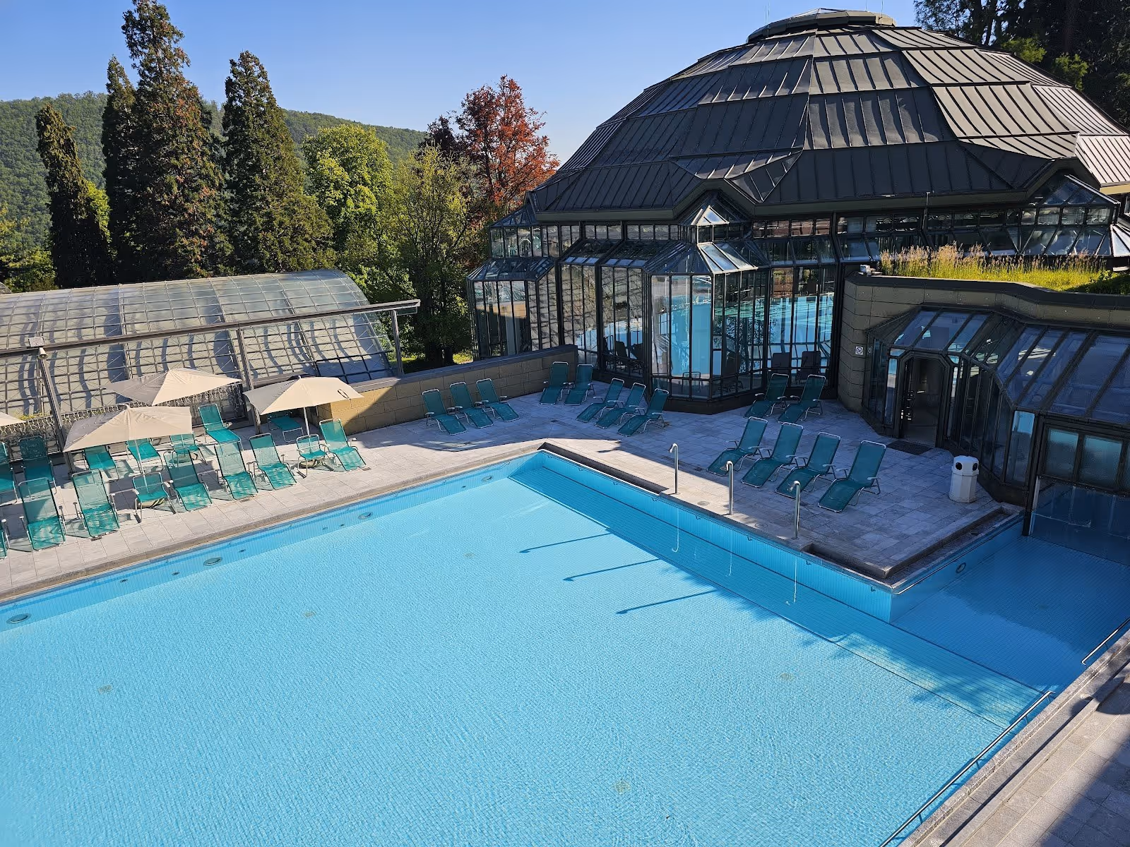 Outdoor swimming pool surrounded by green trees and turquoise lounge chairs at Cassiopeia Therme Badenweiler in Badenweiler, Germany.