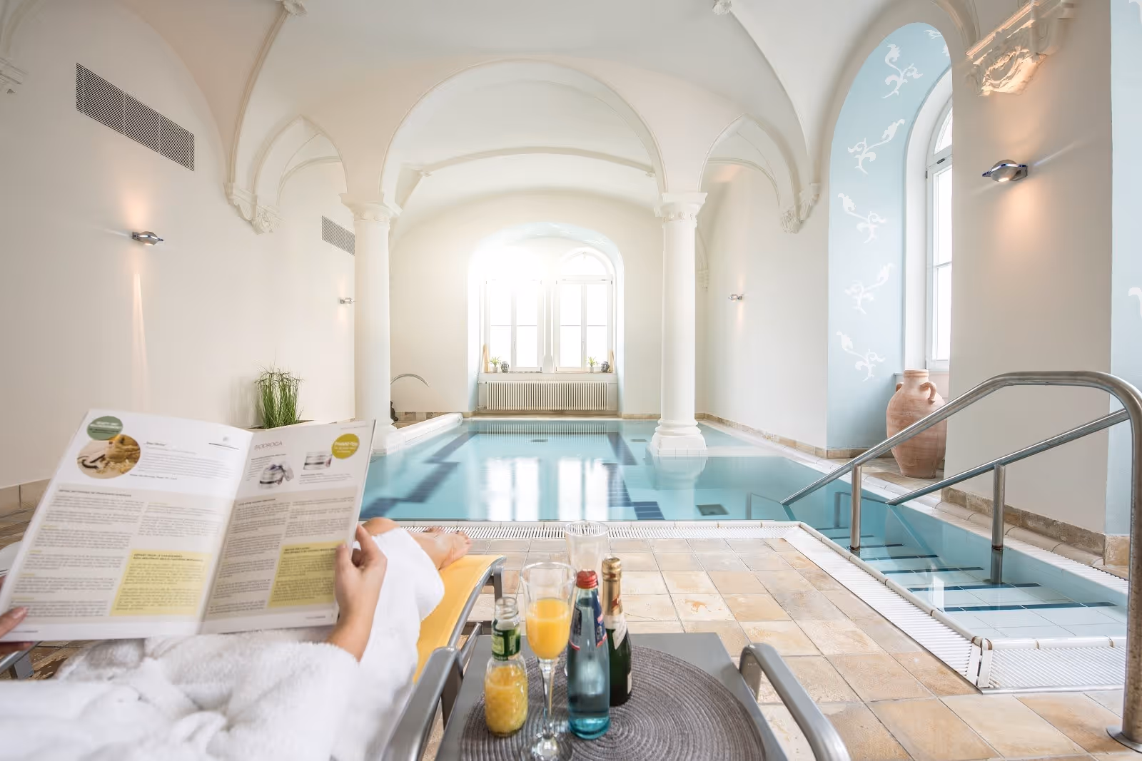 Person relaxing on a lounge chair reading near a bright indoor spa pool with arched windows and white pillars at Friedrichsbad in Baden-Bad.