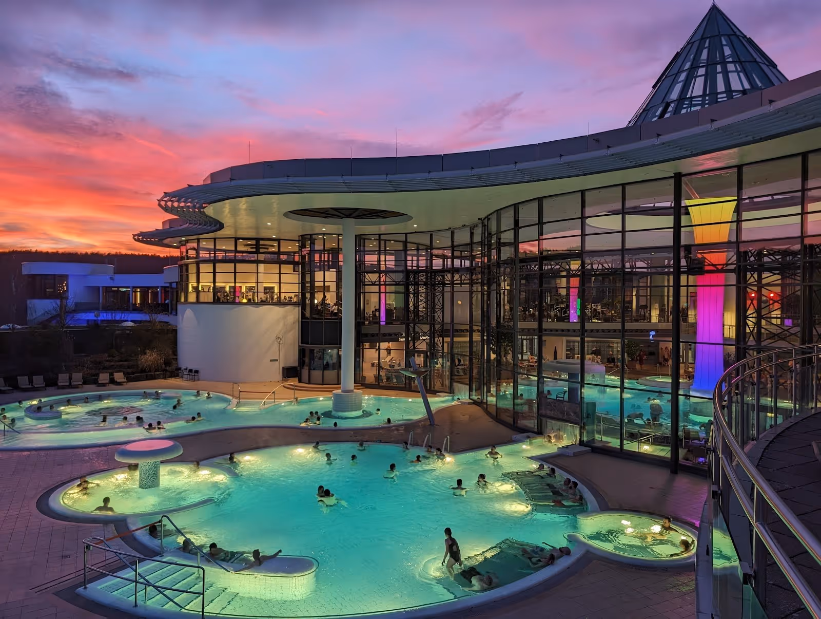 Outdoor hot spring pools at dusk with glowing aqua water and glass building reflecting a colorful sunset at KissSalis Therme in Bad Kissing.