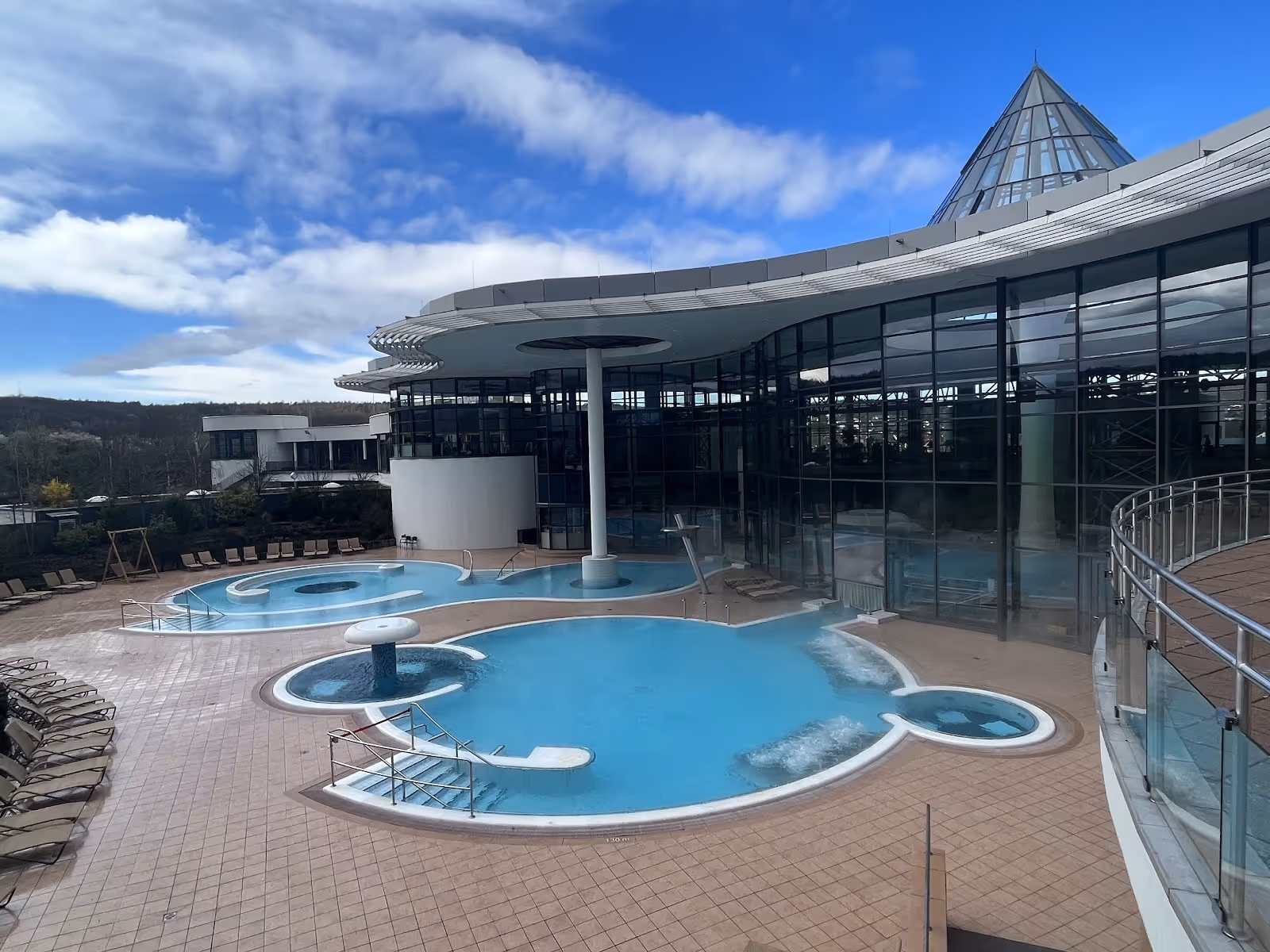 Empty outdoor soaking pools with clean blue water and circular bubbling fountain beside glass walls under partly cloudy sky at KissSalis Th.