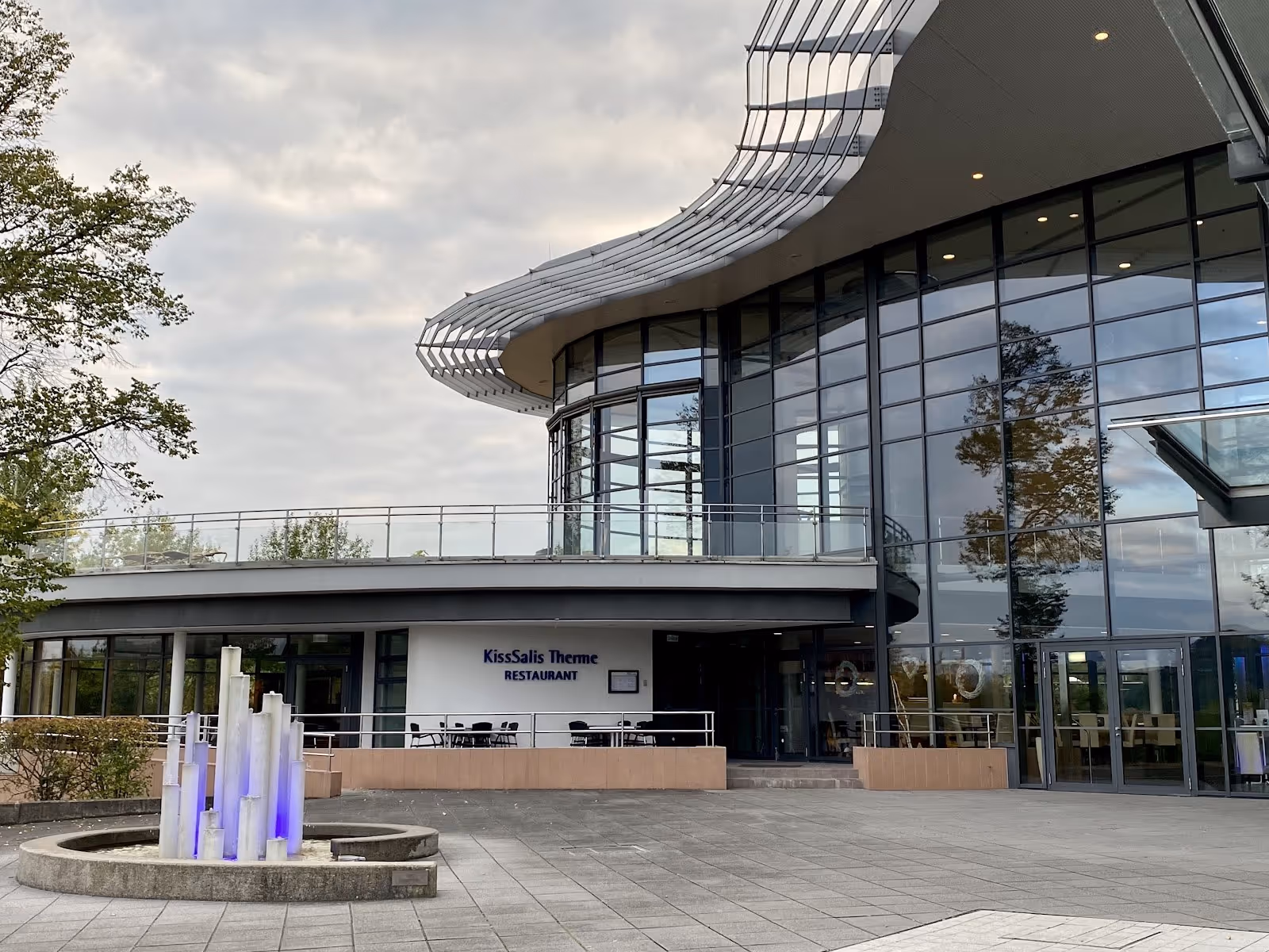 Facade of KissSalis Therme restaurant with tall glass windows and modern roof design next to stone plaza and decorative water feature in Ba.