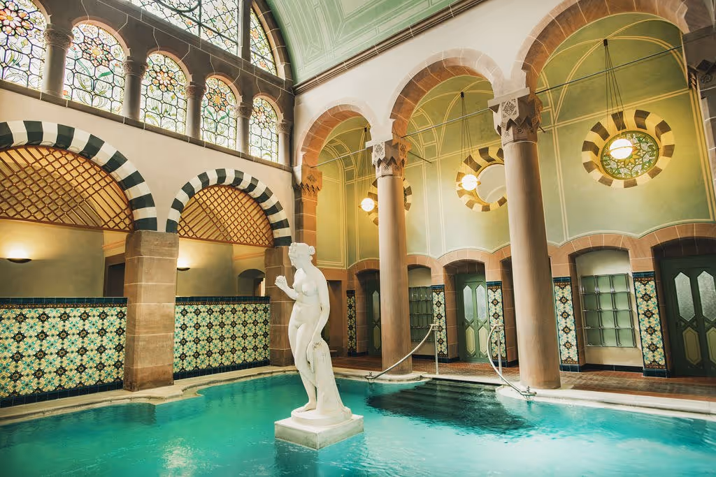 Indoor hot spring pool with turquoise water at Palais Thermal featuring a white statue and green stained glass windows in Bad Wildbad Germa.