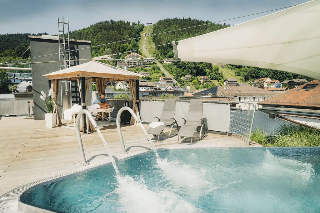 Outdoor rooftop hot spring pool with water jets, lounge chairs, and mountain view overlooking the town of Bad Wildbad at Palais Thermal Ger.