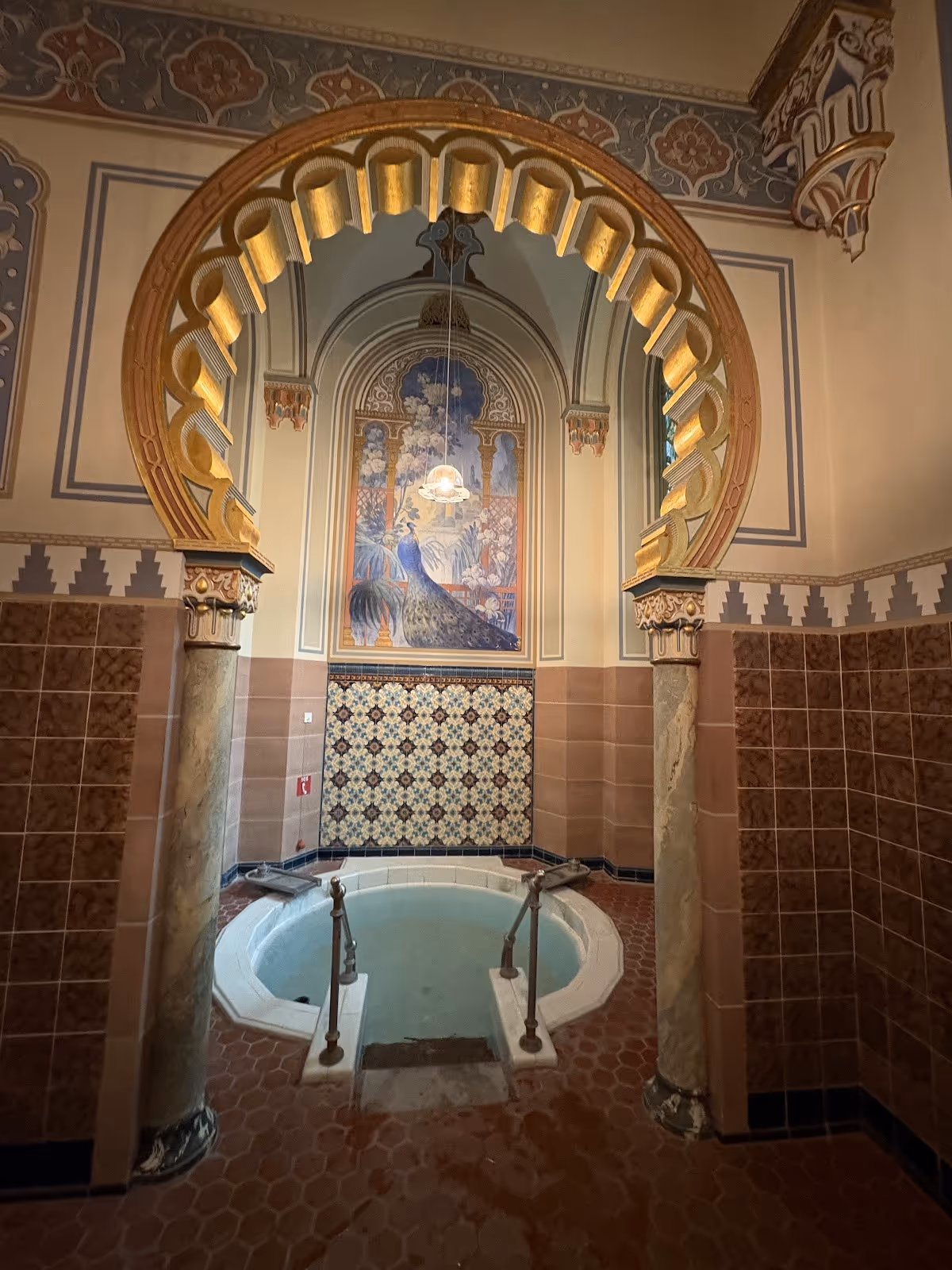 Small indoor hot spring soaking pool at Palais Thermal with decorative tile and a mural of a peacock under an ornate arched ceiling in Bad.