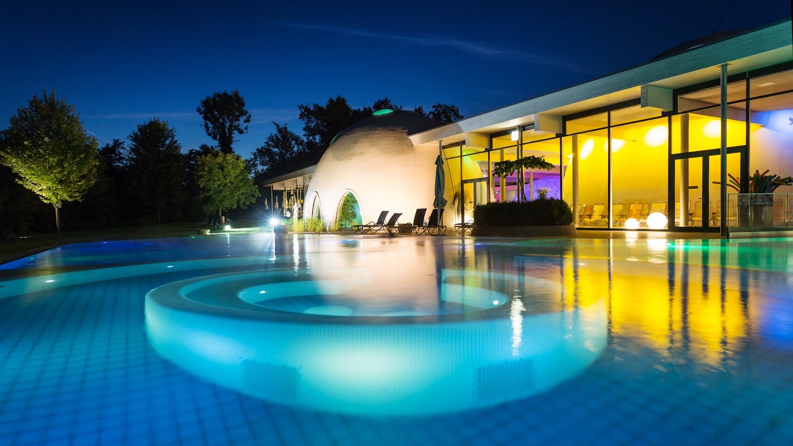 Outdoor spa pool at Therme Bad Aibling in Germany lit with blue and yellow evening lights near curved white dome buildings.