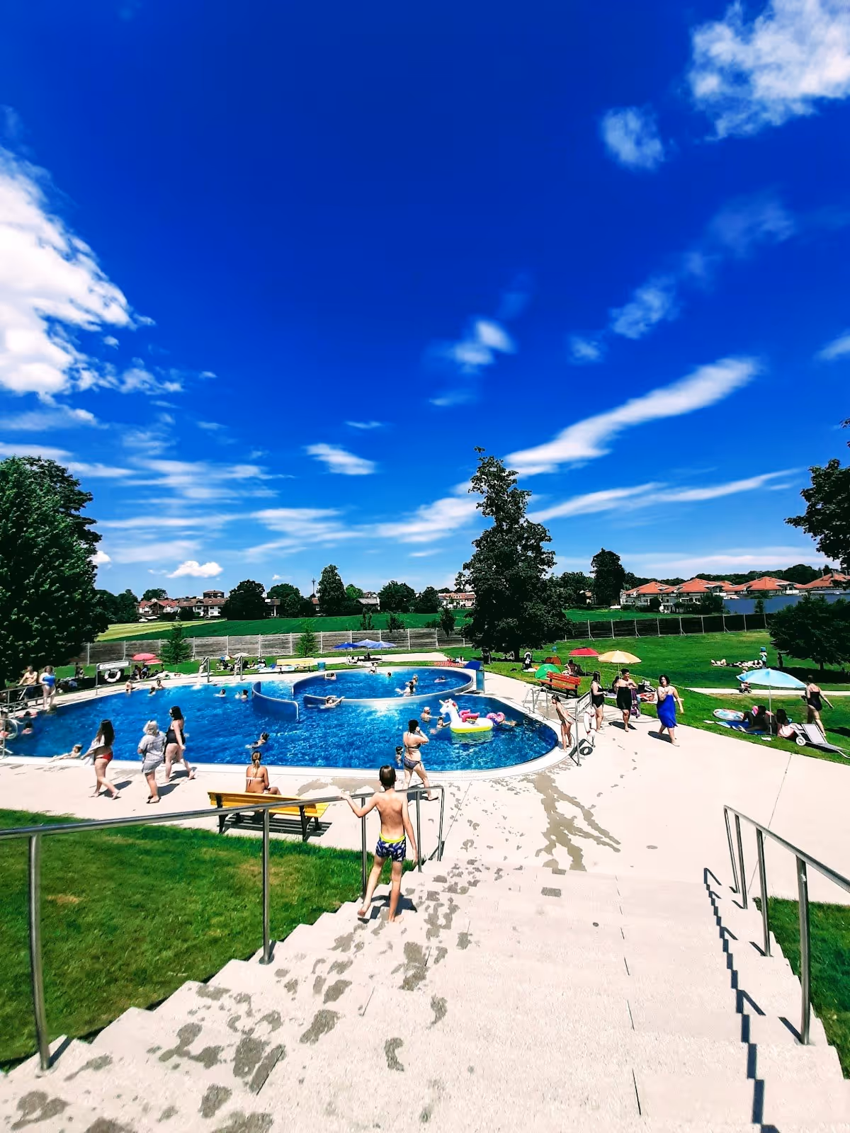 Outdoor public pool facility with people swimming and walking on stairs under blue sky near trees and houses in Bad Aibling, Germany.