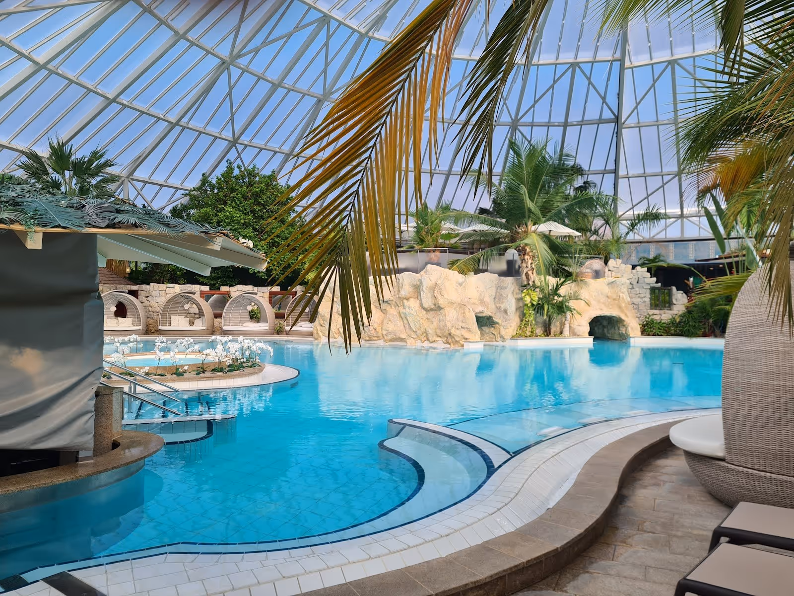 Clear blue water of an indoor mineral pool at Therme Erding, Germany with rock formations and tropical plants visible under glass roof.