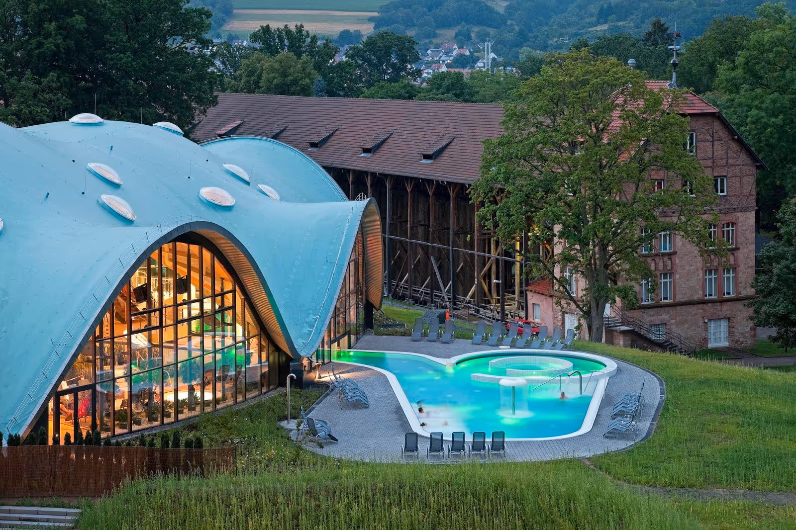 Daytime scene of outdoor mineral pool at Toskana Therme Bad Orb near large curved glass building and green landscape.