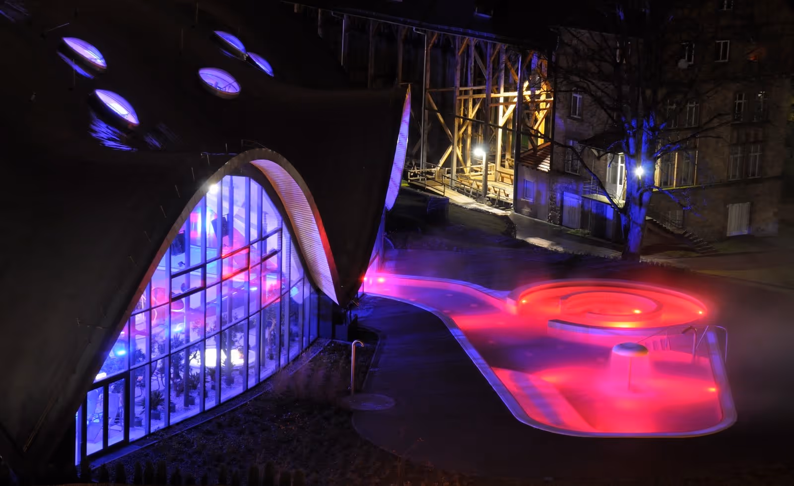 Nighttime photo of Toskana Therme Bad Orb's outdoor soaking pool illuminated with red and purple lights with steam.