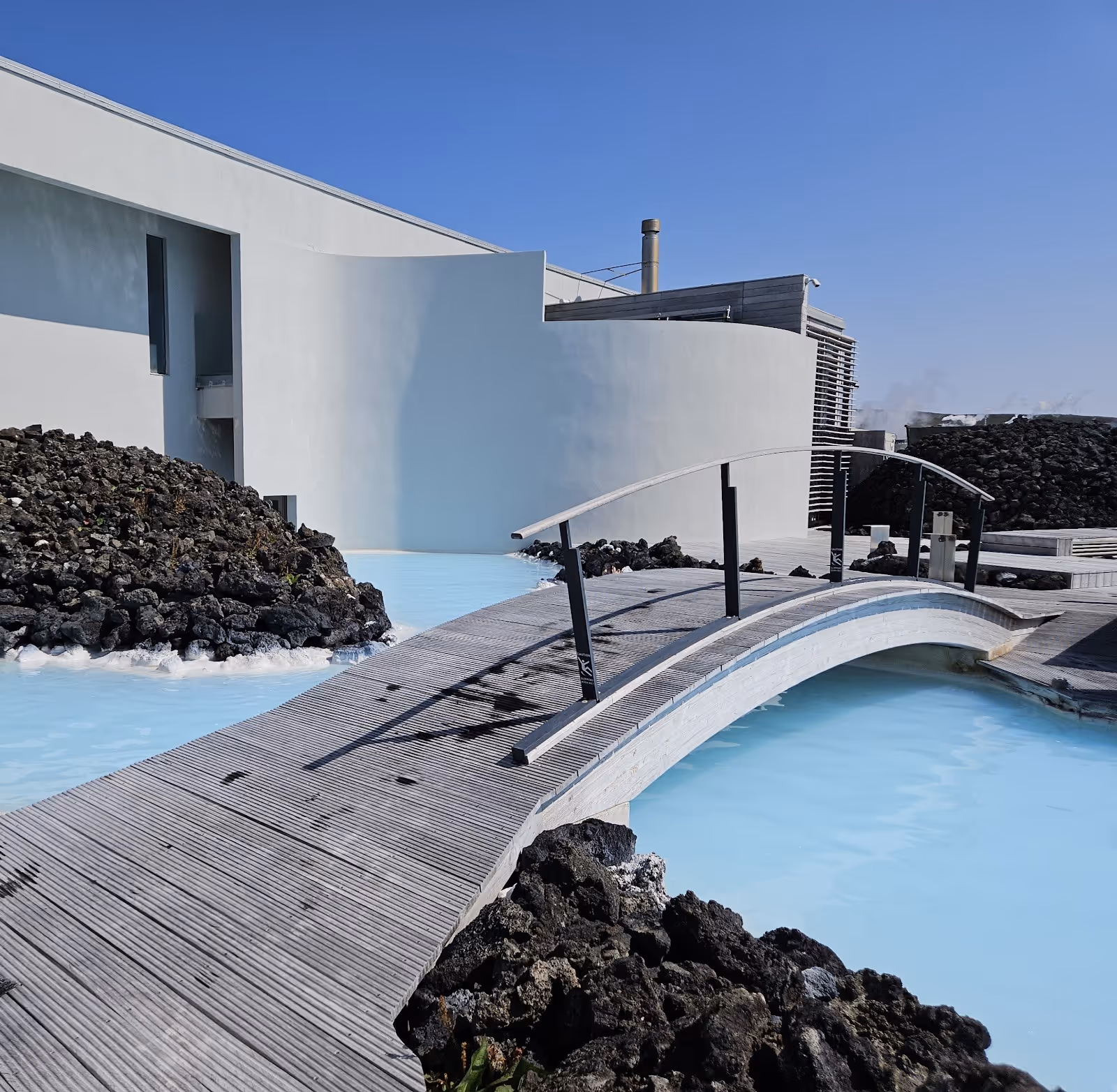 Wooden boardwalk and arched footbridge over bright blue water next to white building and black lava rocks at Blue Lagoon Iceland.