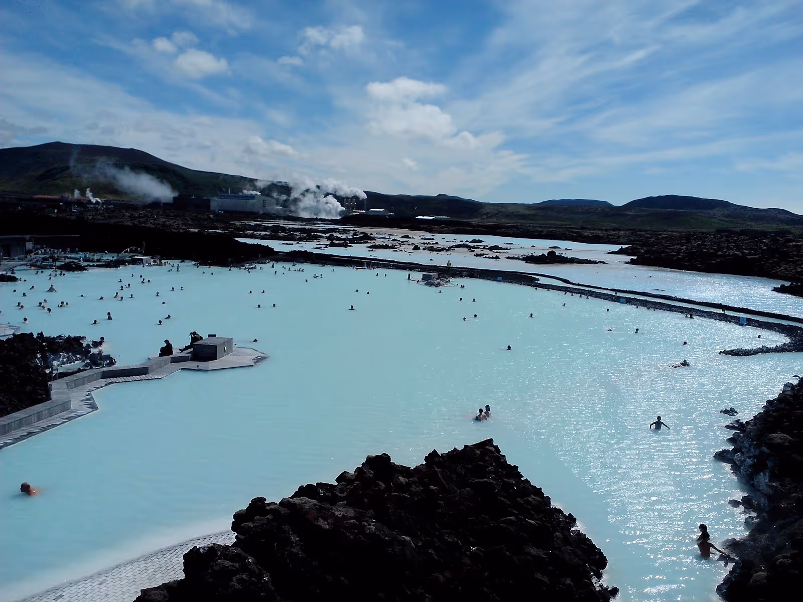 Large Blue Lagoon outdoor mineral pool with distant people soaking and steam rising against Icelandic hills near Grindavik.