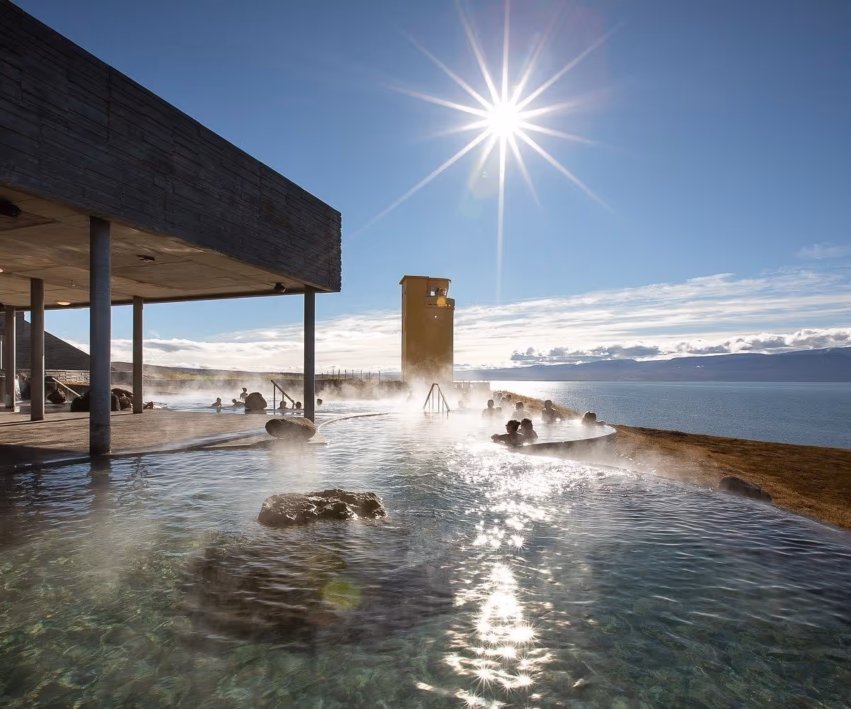 Steam rising from an outdoor hot spring pool with people soaking under bright sun and a yellow tower structure at GeoSea Baths in Iceland.