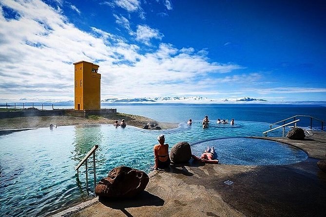 Sun shining over daylight outdoor geothermal pools with people relaxing around a concrete plaza at GeoSea Geothermal Sea Baths in Iceland.