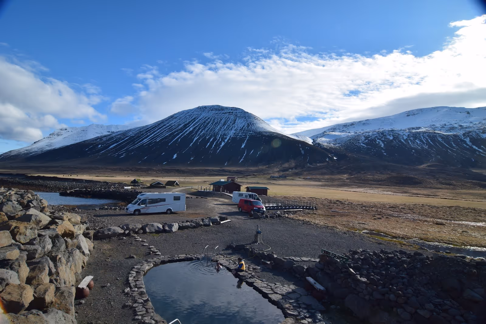 Outdoor hot spring pool next to parked vehicles with snow-capped mountains under a cloudy sky near Saudarkrokur at Grettislaug Hot Springs.