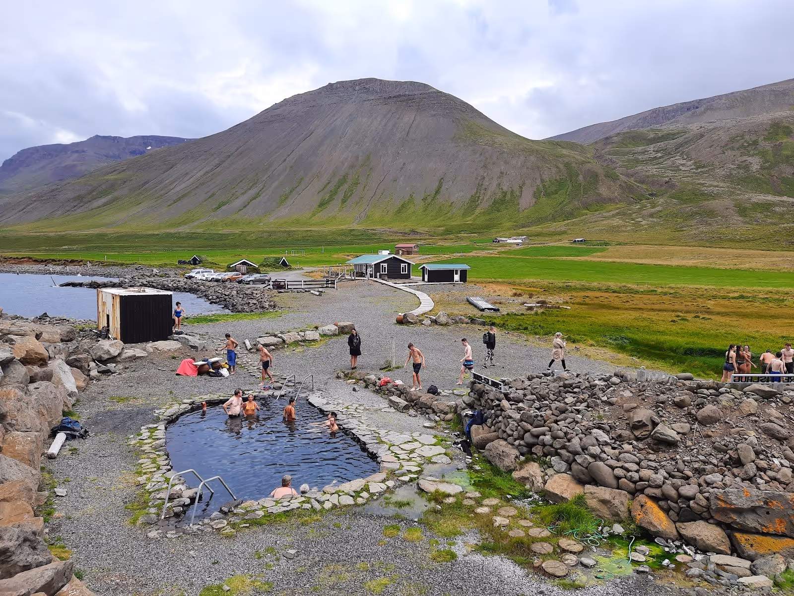 People enjoying Grettislaug Hot Springs soaking pool with green fields and a large mountain in the background near Saudarkrokur Iceland.