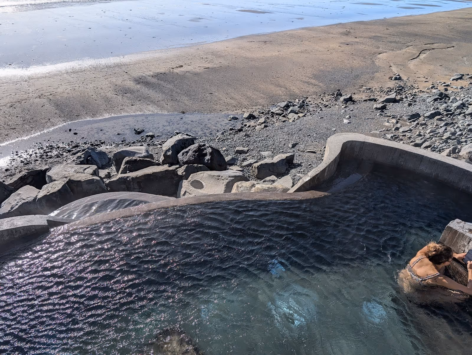Large hot spring pool with rippling water near a rocky shoreline and sandy beach at Gudlaug Baths, Iceland.