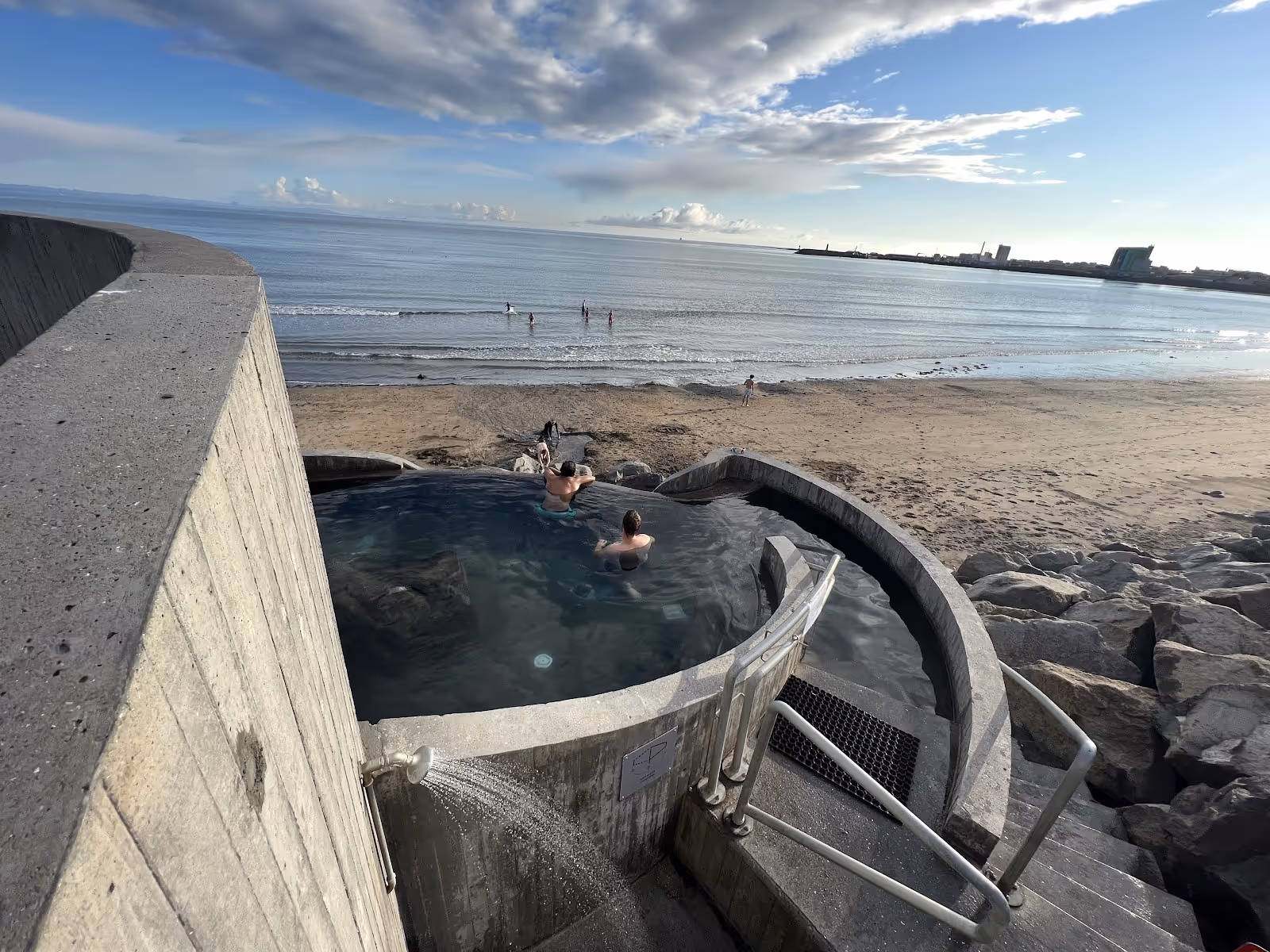 Two people relaxing in a curved concrete hot spring pool overlooking a sandy beach near Akranes, Iceland.