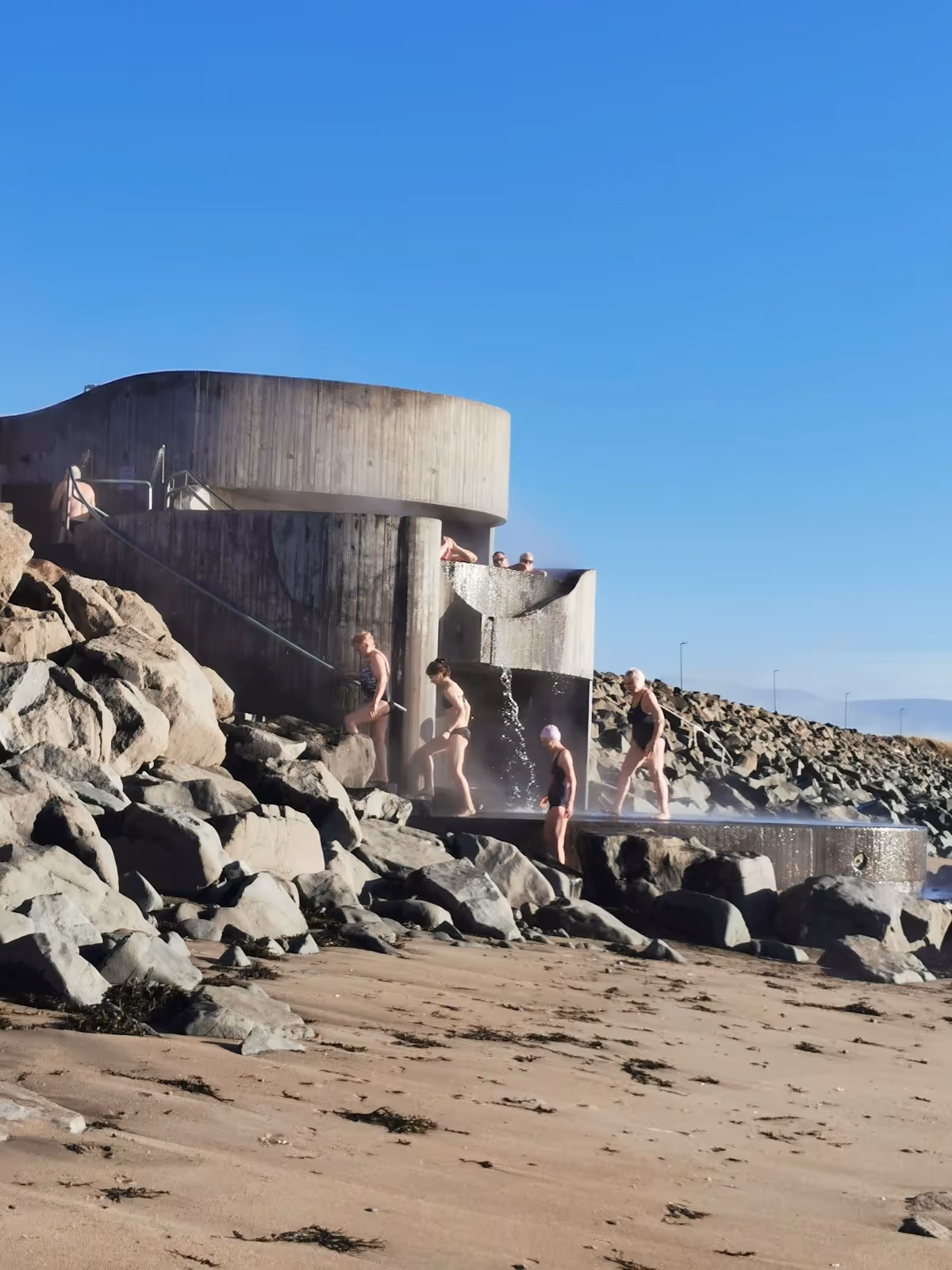Group of people enjoying a hot spring bath next to a spiral concrete structure at Gudlaug Baths in Iceland.