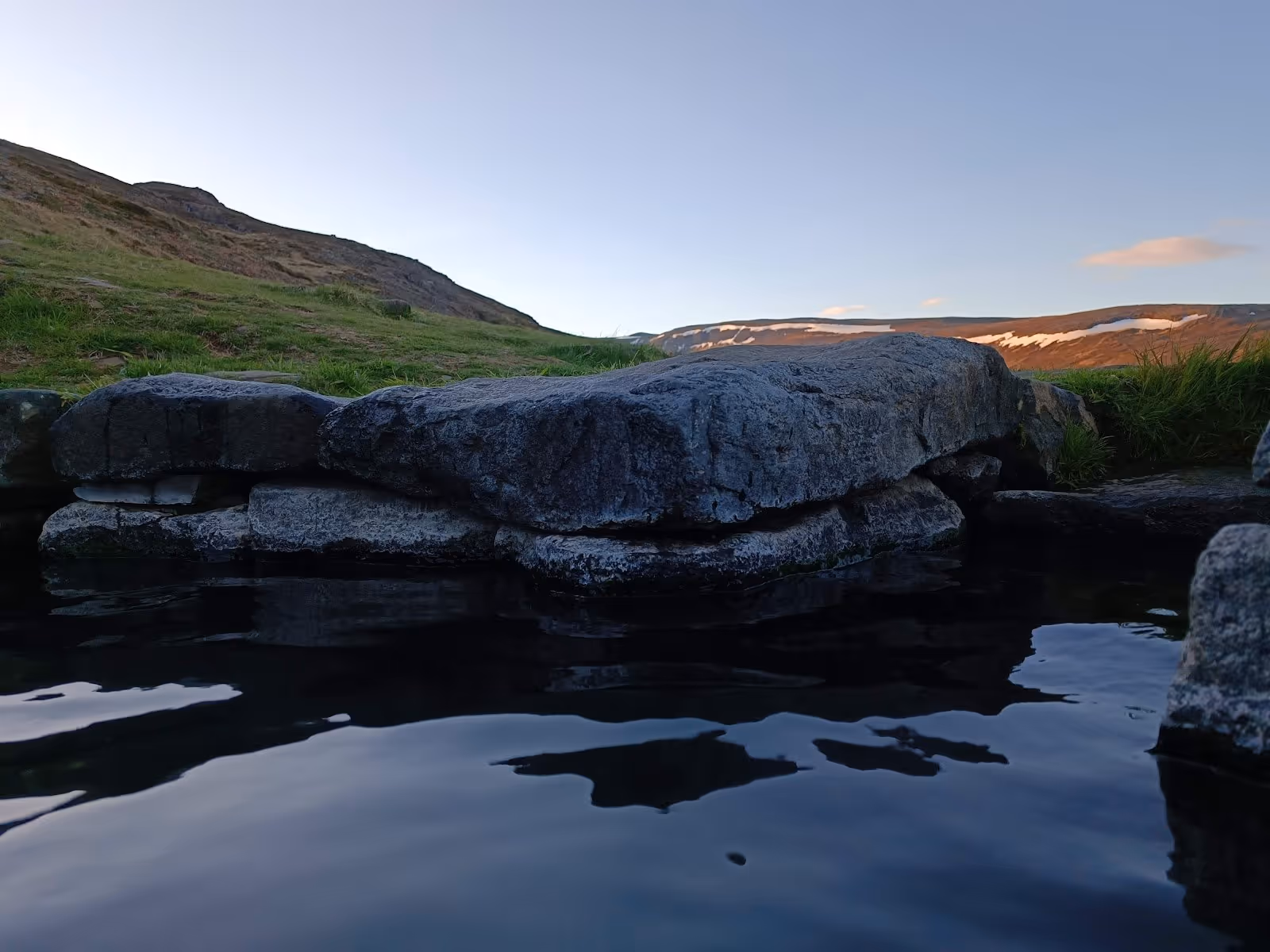 Closeup of dark stone edges surrounding calm water in a hot spring pool at Gudrunarlaug with a green hillside under clear sky.