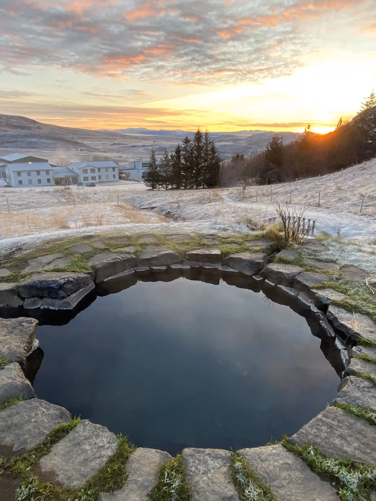 Gudrunarlaug hot spring pool steaming gently at sunset with frosted grass and buildings in the distance in Budardalur Iceland.