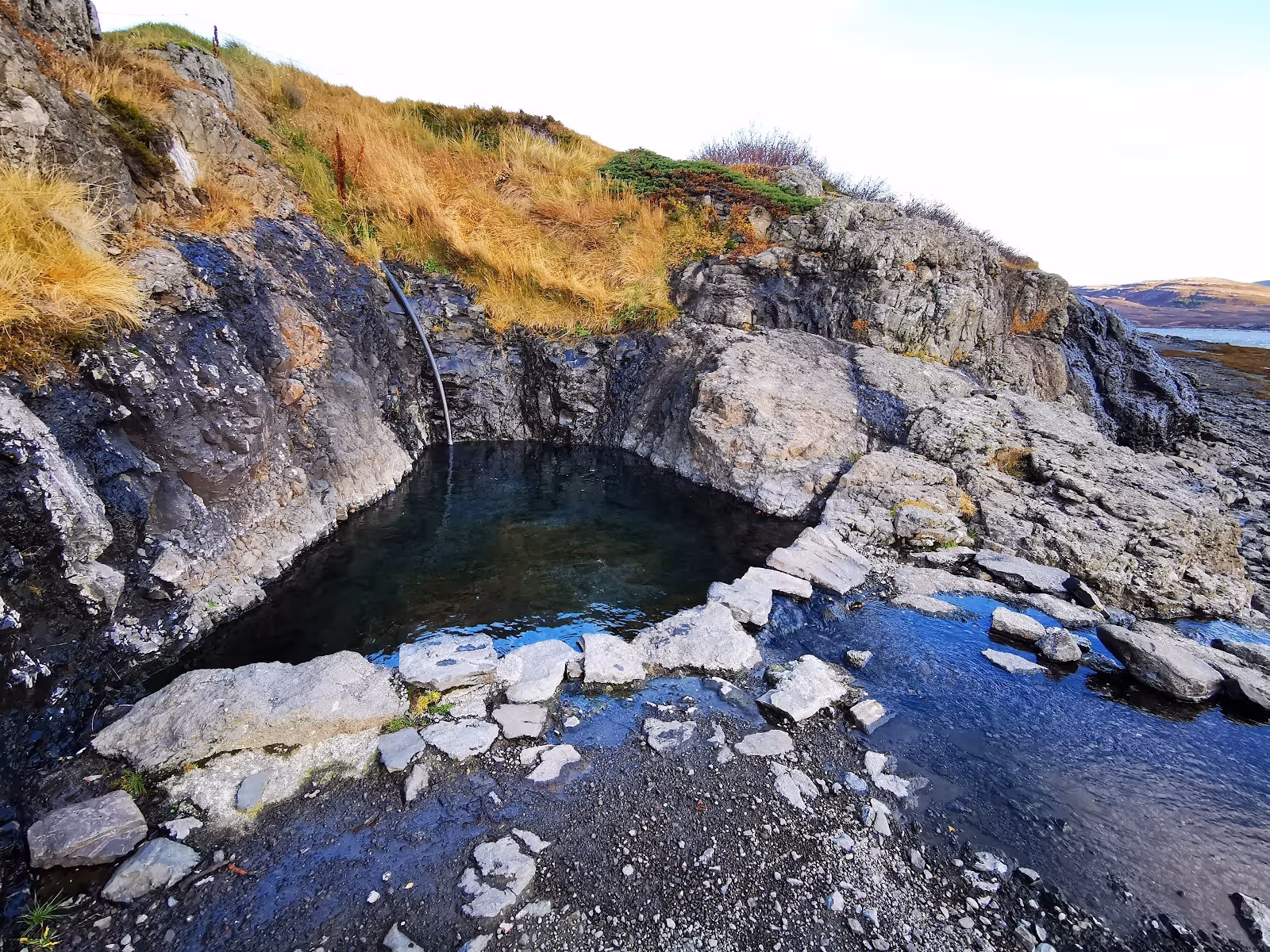 Clear water flowing into Hellulaug Hot Spring pool bordered by stones with grassy hill and rocky coast in Iceland.