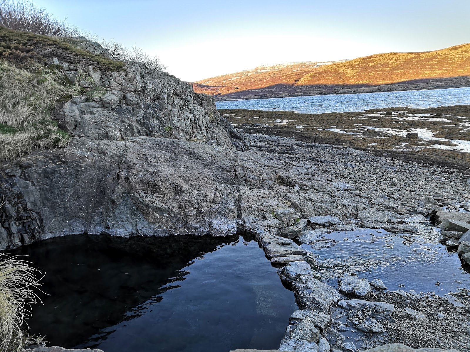 Empty natural hot spring pool carved into rocks at Hellulaug Hot Spring with dry grass and rocky shore near Flokalundur Iceland.