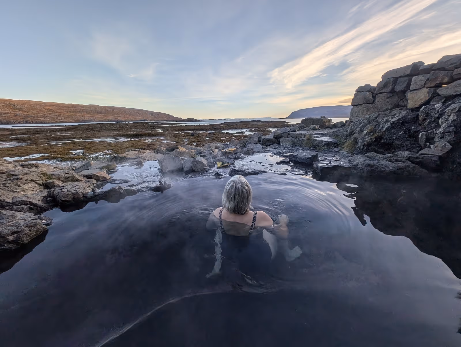 Person soaking in steaming Hellulaug Hot Spring pool with rocky shoreline and distant mountains near Flokalundur Iceland.