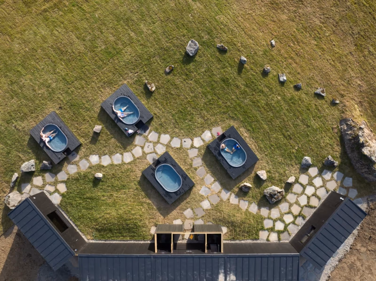 Aerial view of five black-framed tubs with clear water set on a stone path in green grass at Hoffell Hot Tubs Iceland.