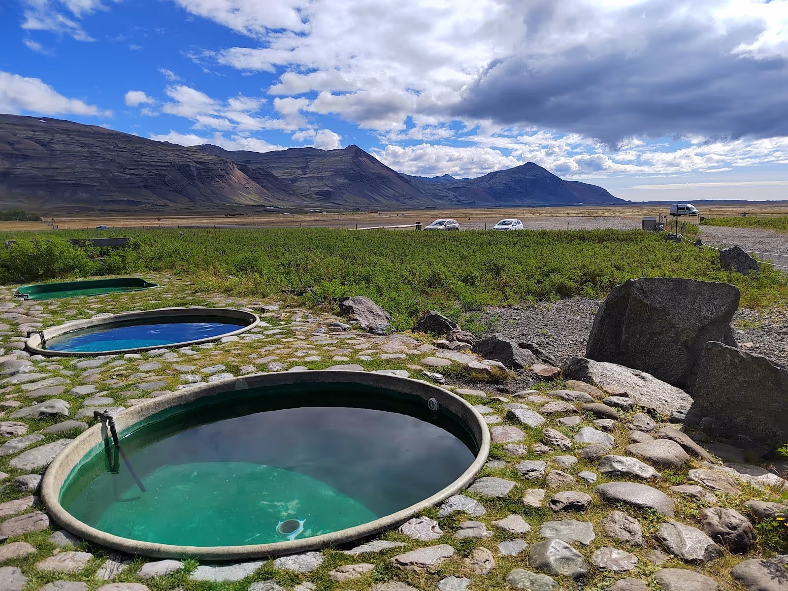 Round mineral pools surrounded by stones steaming slightly with a mountain range near Hofn Iceland in background.