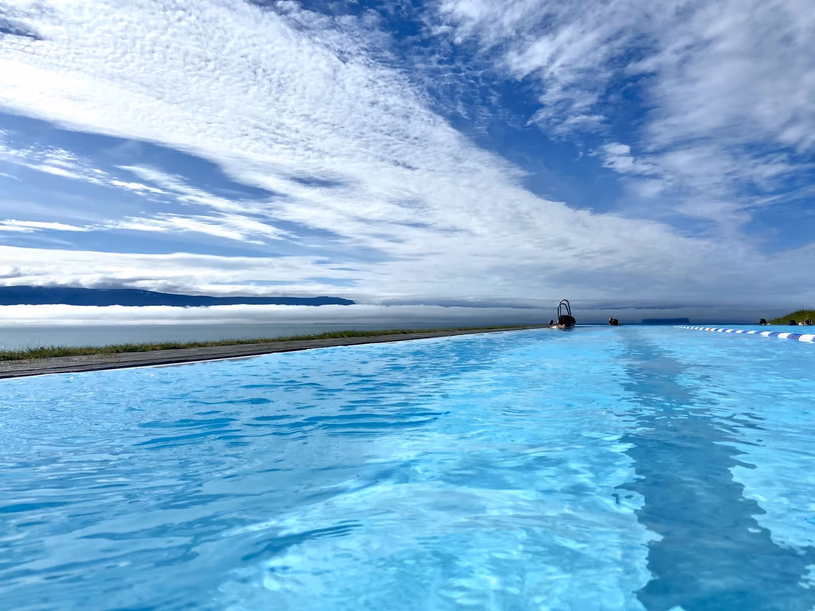 Clear bright blue water in Hofsos Swimming Pool outdoor pool with swimmers at the far end and low fog over water near Hofsos Iceland.