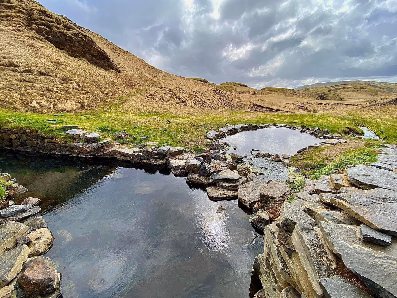 Natural stone hot spring pools with clear water surrounded by grassy hills near Fludir in Iceland at Hrunalaug Hot Spring.