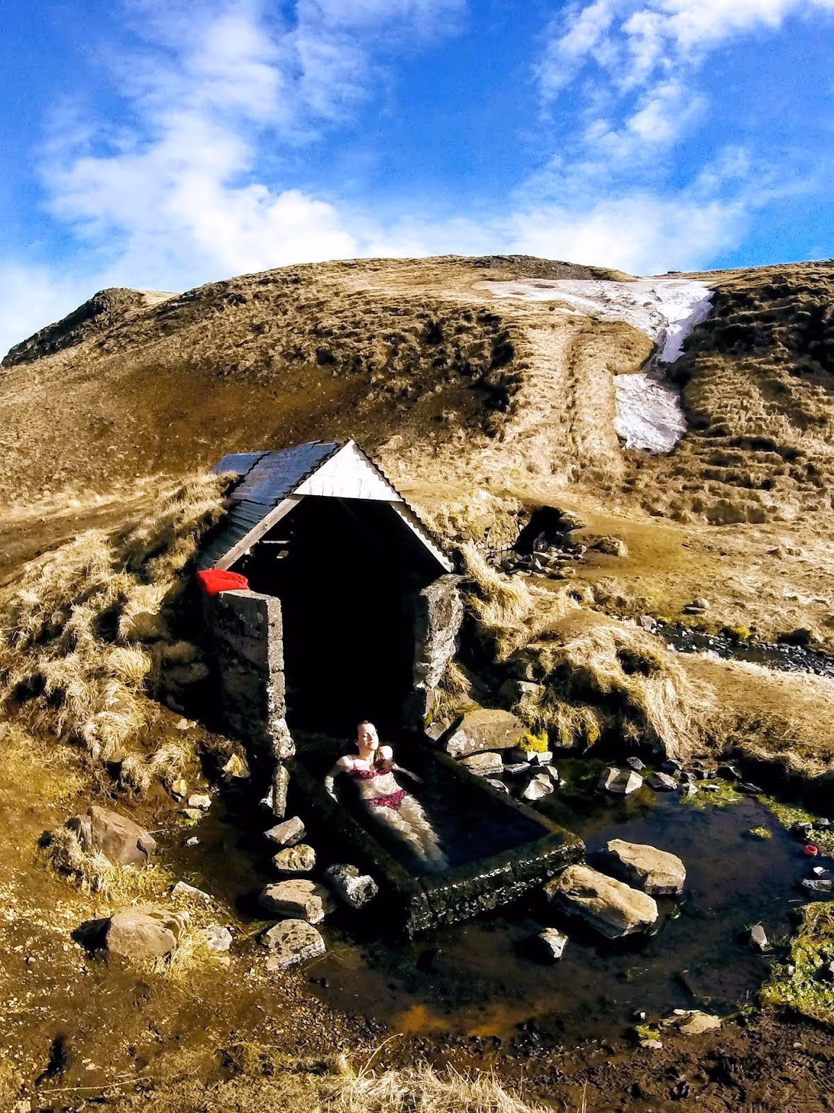 Small outdoor pool with stone walls under a wooden shelter where a person relaxes in Hrunalaug Hot Spring near Fludir, Iceland.