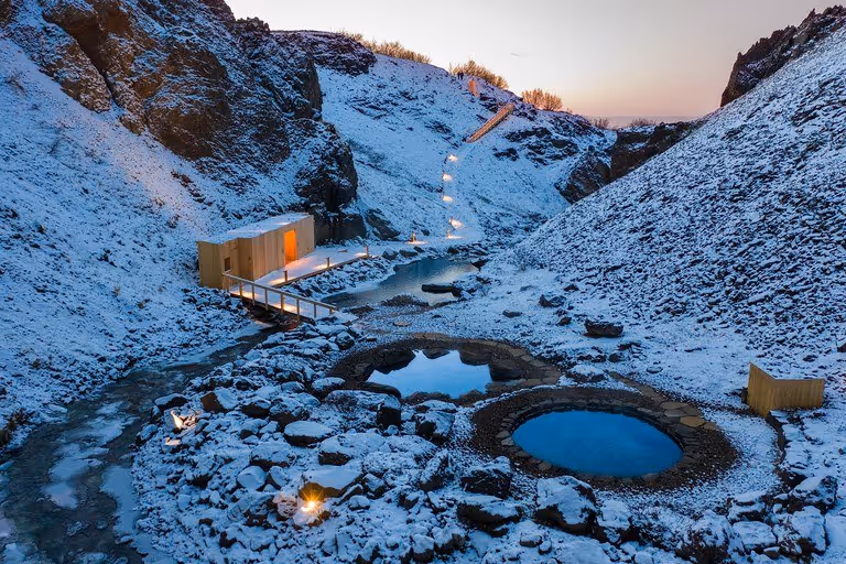Snow-covered rocky hot spring pools connected by wooden boardwalk with glowing lights in Husafell, Iceland canyon setting.