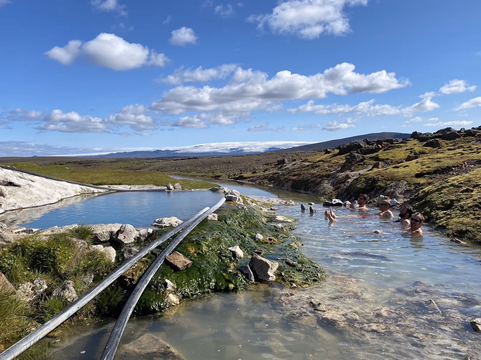 Several people enjoy soaking in narrow natural hot spring pools surrounded by rocks and patches of grass at Hveravellir near Hveravellir, I.