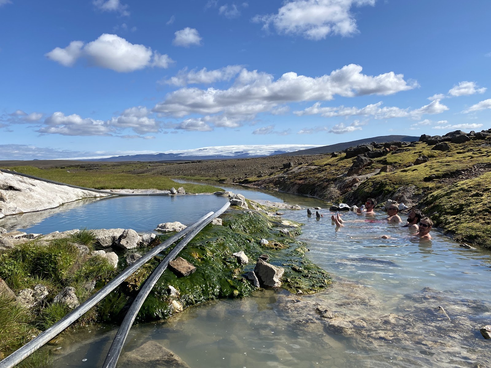 Several people enjoy soaking in narrow natural hot spring pools surrounded by rocks and patches of grass at Hveravellir near Hveravellir, I.